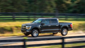 A dark-colored 2021 Ford F-150 travels on a rural dirt road flanked by wooden fencing