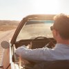 Man wearing old money style clothes while driving a classic convertible on a dirt road.